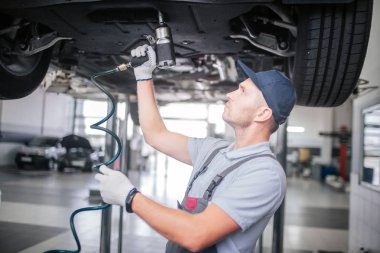 Ayakta ve arabanın altındaki çalışma younf adam resmi. O arama yapar. Adam tutar ve matkap kullanır. O beyaz eldiven çalışıyor