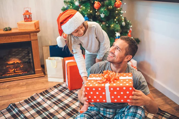 Père et fille joyeux se regardent et sourient. Il est assis sur une couverture par terre et regarde sa fille. Elle l'a regardé en bas. La fille porte un chapeau. Il y a cheminée et arbre de Noël derrière . — Photo