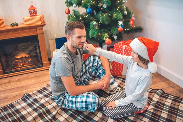 Preciosa foto de una chica tocando la nariz de su padre con el dedo. Ella lo mira y sonríe. Un joven en pijama mira a su hija. Él también sonríe. Se sientan en la habitación decorada . — Foto de Stock