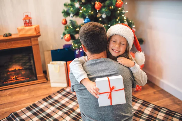 Feliz niña sonriendo. Ella recibió un regalo de su padre. El chico lo abraza. Ella es feliz. La chica lleva sombrero. Están en la habitación con chimenea y árbol de Navidad . — Foto de Stock