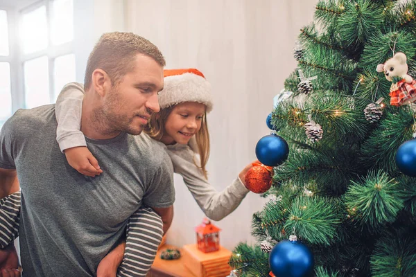 Photo de fille assise sur les papas dos et décorer l'arbre de Noël avec des jouets. Elle y met un jouet rouge. Jeune homme la tient très précise . — Photo