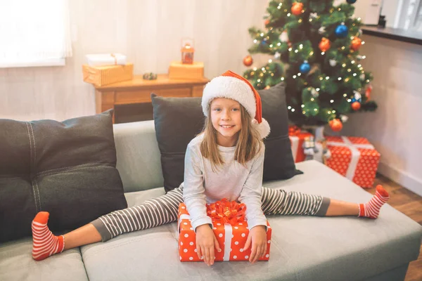 Small and happy girl posing on camera. She looks happy. Girl sits on twine and leans on red box with present. She looks on camera. Girl is alone in room.