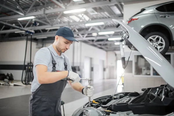 Młodego pracownika stoi otwarty ciało samochodu. Używa wrech, aby naprawić. On jest poważne i skoncentrowany. Facet pracuje w garażu. — Zdjęcie stockowe