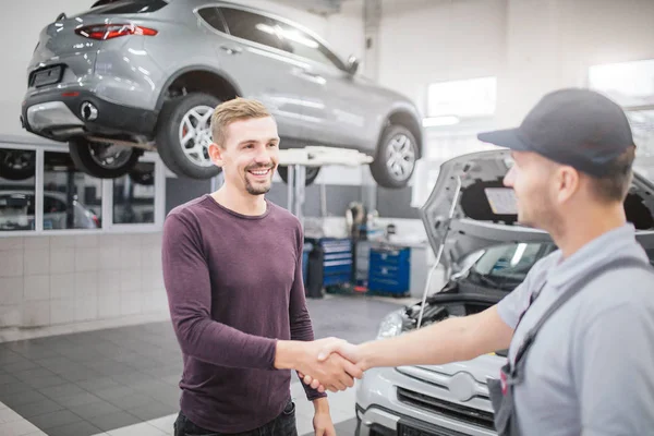 Gli uomini felici stanno in garage e si stringono la mano. Si trovano di fronte ad una carrozzeria aperta. C'è un altro veicolo dietro l'uomo biondo sulla piattaforma . — Foto Stock