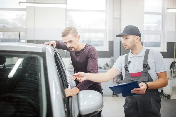 Dos jóvenes serios están en el auto. Propietario se inclina a ella y mira dentro del salón. Puntos de trabajo con pluma. Está concentrado. También chico en uniforme sostiene tableta de plástico con papeles . — Foto de Stock