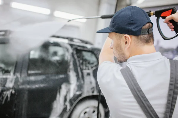 Trabajador en pie uniforme gris en coche negro cubierto con espuma. Él sostiene la manguera flexible con la pistola en él y lavar el automóvil. El hombre es serio y concentrado. Está en el garaje. . — Foto de Stock