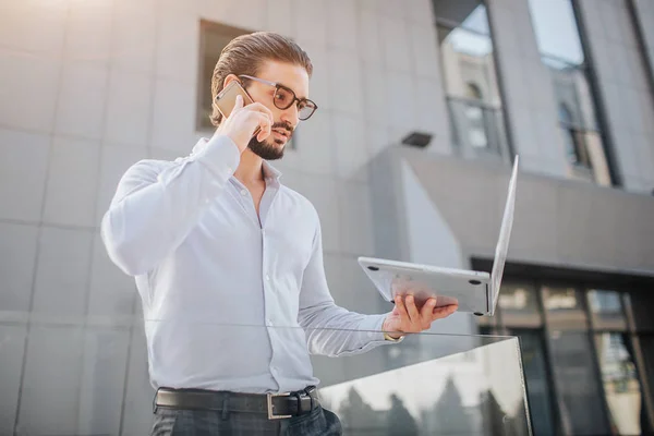 Concentrated and handsome young businessman stands and poses. He looks at laptop screen and talks on phone at the same time. There is building behind him. — Stock Photo, Image