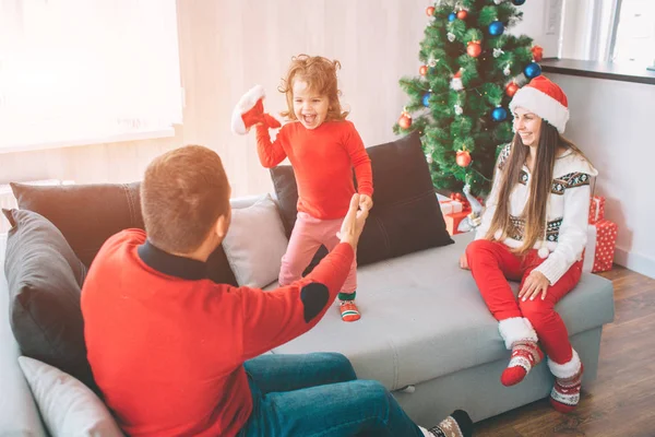 Joyeux Noël et bonne année. Photo ludique d'un enfant heureux debout sur un canapé et tenant un chapeau rouge. Elle regarde son père et crie. Le jeune père tient ses filles par la main. Look femme et sourires . — Photo