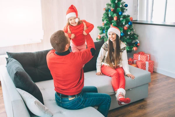 Joyeux Noël et bonne année. Charmante image de la famille heureuse assise sur le canapé. Papa joue avec un gosse. Il tient la fille dans ses mains. Elle sourit. L'enfant est heureux. Elle porte un chapeau. Une femme les regarde. — Photo