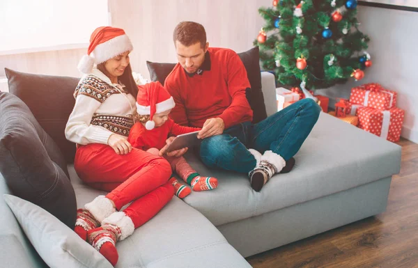 Joyeux Noël et bonne année. Famille dans des vêtements chauds et chapeaux assis ensemble sur le canapé. Ils regardent. Fille tient la tablette dans les mains. Aidez-la. Ils sont concentrés . — Photo