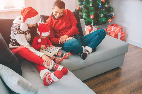 Joyeux Noël et bonne année. Famille assise ensemble sur le canapé. Une petite fille est entre ses parents. Elle tient et regarde le livre avec des photos. L'homme le regarde et sourit. Ils sont heureux. . — Photo