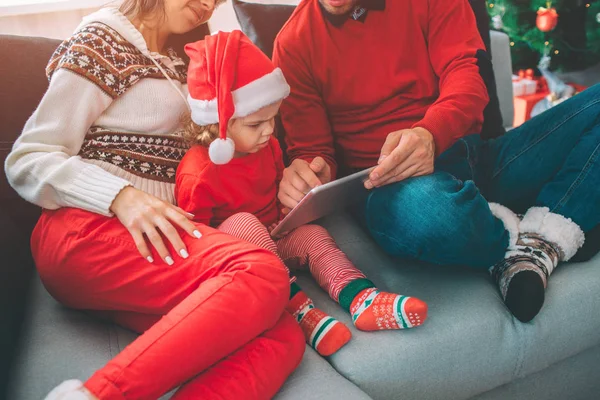 Merry Christmas and Happy New Year. Family Cut view of small girl sits between her parents. She holds tablet and points on it. Girl touches screen with finger. Man help to hold tablet. — Stock Photo, Image