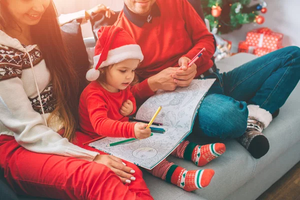 Joyeux Noël et bonne année. Couper la vue des parents assis sur le canapé avec leur enfant. Le gamin tient de la coloration et des crayons dessus. Fille dessiner avec elle. Le jeune homme tient aussi des crayons. . — Photo