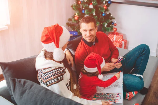 Joyeux Noël et bonne année. Belle image de la famille assise ensemble sur le canapé. Les parents se regardent. Une femme porte un chapeau. Le jeune homme sourit. Leur fille dessine dans la coloration . — Photo