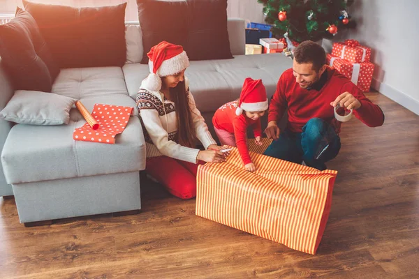 Joyeux Noël et bonne année. Photo d'un petit assistant tenant un morceau de papier pour cadeau. Ses parents sourient. Le jeune homme tient du ruban adhésif et l'utilise pour couvrir le présent. Femme les aide . — Photo