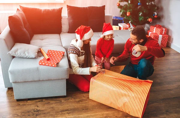 Joyeux Noël et bonne année. Famille assis sur le sol près de la grande boîte de cadeau. Le jeune homme montre à sa fille comment coller le bord de la boîte. La fille regarde ça. Elle est concentrée. Femme regarde fille . — Photo