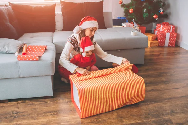 Joyeux Noël et bonne année. Jeune femme et petite fille s'assoient ensemble. Enfant sur les genoux des mères. Ils préparent la couverture pour le présent. L'enfant l'aide. Ils travaillent en équipe. . — Photo