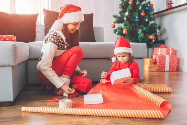 Frohe Weihnachten und ein gutes neues Jahr. kleines Mädchen sitzt auf dem Boden. Sie hält eine weiße Schachtel in den Händen. Kind versucht, es mit Weihnachtsdeckel zu packen. junge Frau lächelt. Sie schaut sich Prozess an. — Stockfoto