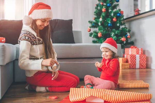 Joyeux Noël et bonne année Une jeune femme et une jeune fille positives et ludiques s'assoient par terre. Ils sourient et rient. L'enfant tient une partie de la bande pendant que la femme se repose. Ils portent des chapeaux. Les filles préparent des cadeaux . — Photo