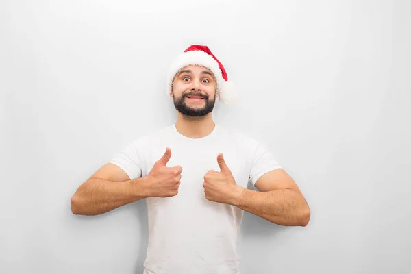 Positieve jonge man blikt op camera. Hij toont zijn grote duimen omhoog. Guy draagt Chritmas hoed. Geïsoleerd op witte achtergrond. — Stockfoto