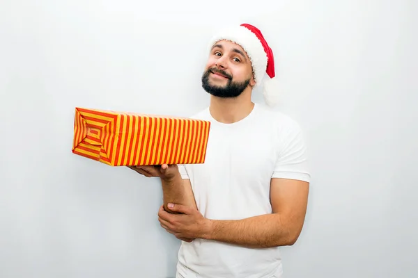 Nice bearded man in red hat stands and holds orange box with present in one hand. He looks on camera and smiles a bit Isolated on white background. — Stock Photo, Image