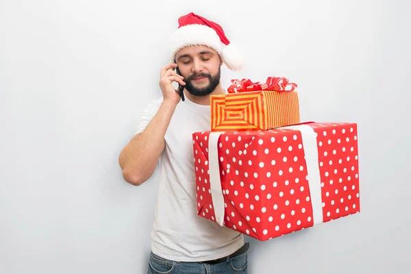 Busy young man stands and talks on phone. He holds bog red box with ribbons and present inside it. Guy looks down. Isolated on white background. — Stock Photo, Image