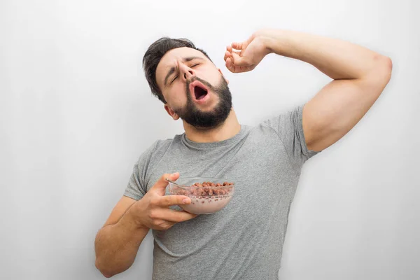 Sleepy male person stands and stretches his hand. He holds bowl with milk and chocolate chunch breakfast. Isolated on white background.