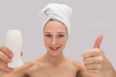 Olumlu ve neşeli genç kadın anlamına gelir ve kamera üzerinde görünüyor. O onun büyük başparmak ve deodorant başka bir elinde tutar. Saçlarını etrafında beyaz havlu vardır. Gri arka plan üzerinde izole.