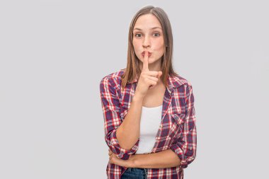 Ciddi genç kadın parmağını dudaklarına tutar. Başka bir el HIPS üzerinde olsun. Kamera için düz görünüyor. Gri arka plan üzerinde izole.