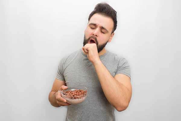 Slaperige oyung man geeuw. Hij hiervoor mond met de hand. Guy heeft ook kommetje melk vol met chocolade crunches. Geïsoleerd op witte achtergrond. — Stockfoto