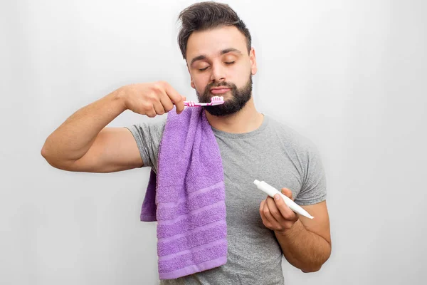 Oicture av sömnig ung man håller tandborste i ena handen och tand klistra in i ett annat. Han håller slutna ögon. Killen har även handduk över axeln. Isolerad på vit bakgrund. — Stockfoto