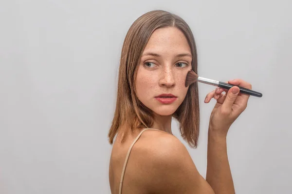 Belle et gentille jeune femme se tient debout et pose. Elle se maquille avec un pinceau sur ses poussins. Elle regarde à gauche et se retourne derrière. Isolé sur fond gris . — Photo