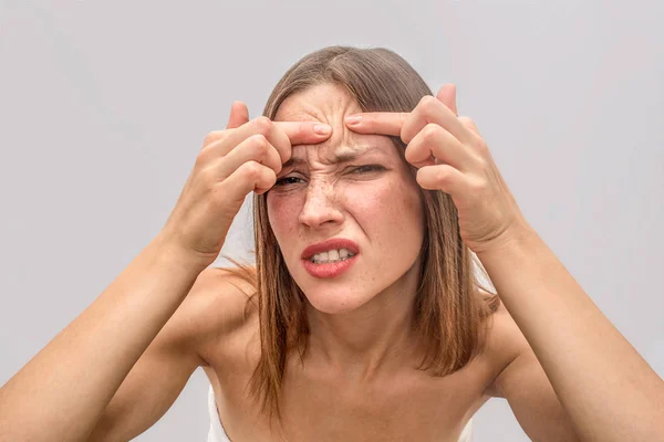 Jovem irritada se levanta e olha mais perto da câmera. Ela mantém os dedos juntos e tenta remover o ponto achne na testa. Isolado sobre fundo cinzento . — Fotografia de Stock