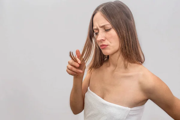 Unhappy and unsatisfied young woman stands and looks at her hair. She touches her hair and looks at it. Woman wears white towel across her body. Isolated on grey background. — Stock Photo, Image