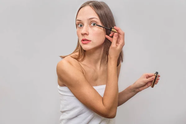 Femme blonde mince et bien construite se tient debout et regarde la caméra. Elle utilise du mascara pour les cils. Elle est concentrée et calme. Isolé sur fond gris . — Photo