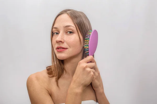 Beau et jeune modèle se tient et regarde la caméra. Elle sourit un peu. Jeune femme tient une brosse et l'utilise pour ses cheveux. Elle est ravissante. Isolé sur fond gris . — Photo