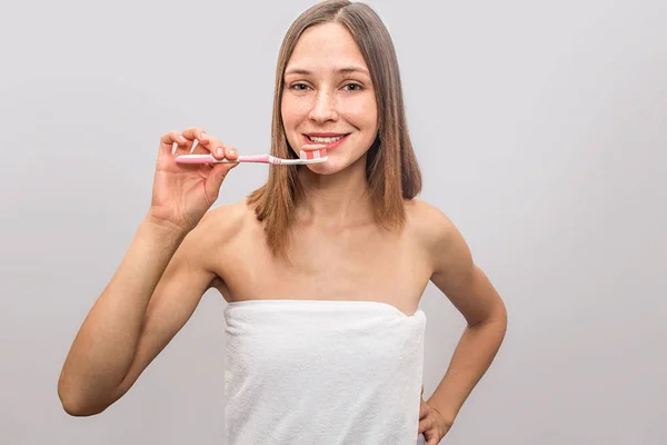 Joyeux et agréable jeune femme se tient debout et pose sur la caméra. Il tient la brosse à dents près de la bouche. Un sourire de mannequin. Une serviette blanche recouvre son corps. Isolé sur fond gris . — Photo