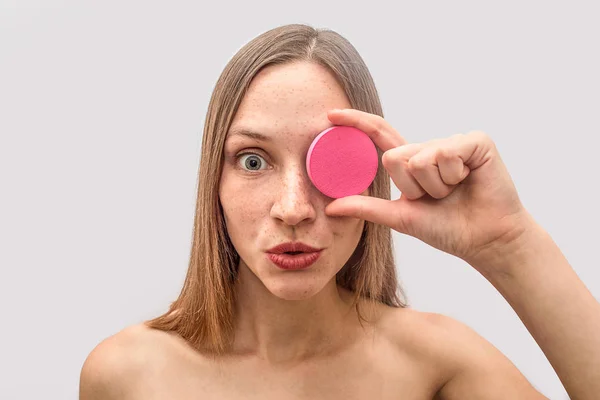 Jeune femme blonde tient spogne rose et couvre les yeux avec elle. Elle est étonnée et s'interroge. Le mannequin regarde la caméra. Isolé sur fond gris . — Photo