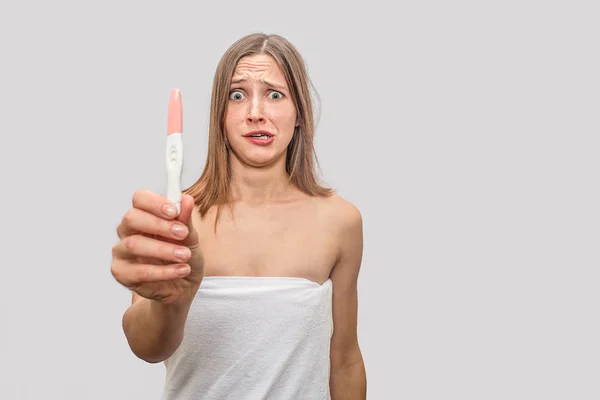 Distracted and unhappy young woman stands and holds pregnancy test. It shows positive result. Model doesnt know what to do. Isolated on grey background. — Stock Photo, Image