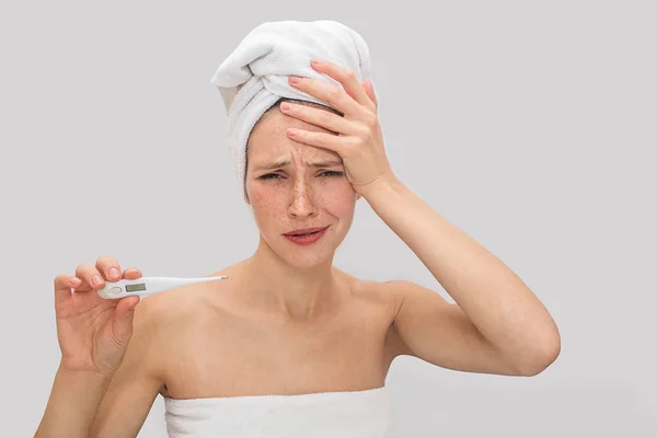 Une jeune femme énervée et triste tient la main sur le front. Elle a de la température. Une jeune femme tient un thermomètre dans la main droite. Elle est malade. Isolé sur fond gris . — Photo