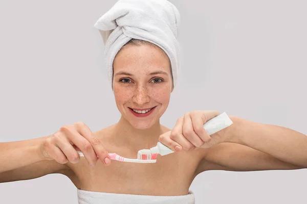 Une jeune femme aux taches de rousseur positive regarde la caméra et sourit. Elle met du dentifrice sur une brosse à dents. Modèle a des serviettes blanches autour de son corps et les cheveux. Isolé sur fond gris . — Photo