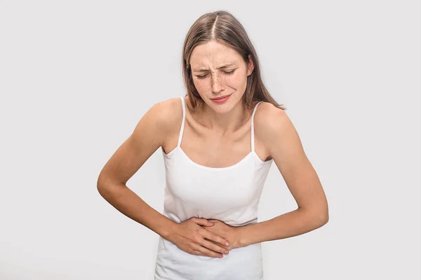 Blonde young woman with freckles stands and holds hands on stomach. She keeps eyes closed. Model suffering. Isolated on grey background. — Stock Photo, Image