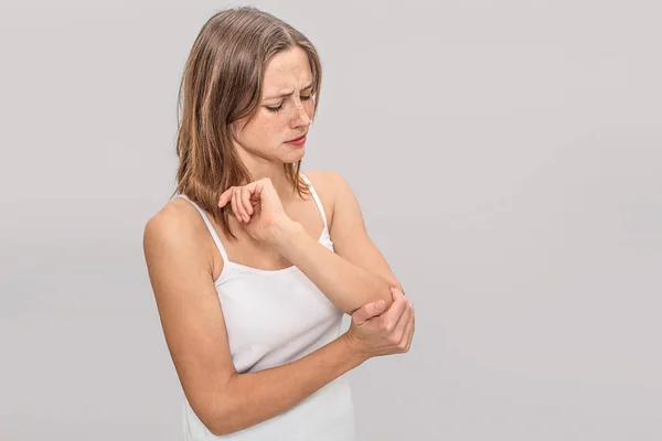 Young woman with freckles holds hand on elbow. There is pain. She is serious. Young woman wears white t-shirt. Isolated on grey background. — Stock Photo, Image