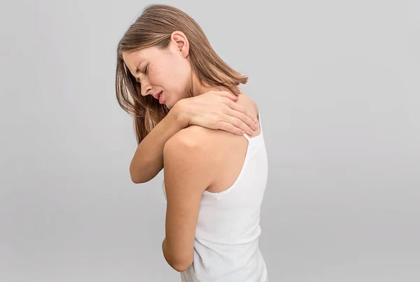 Young and slim woman stands back and holds hand on it. There is pain. She suffers. Model keeps eyes closed or look down. She is exhausted. Isolated on grey background. — Stock Photo, Image