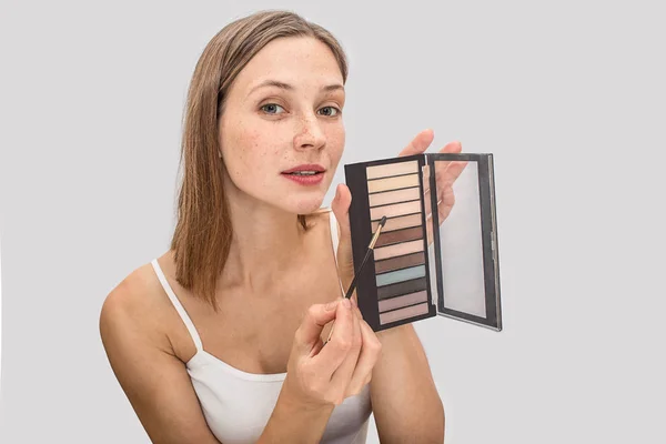 Photo de jeune modèle avec des taches de rousseur sur le visage regardant la caméra et posant. Elle tient une palette d'ombres à paupières et pointe sur une couleur avec un pinceau. La jeune femme est ravissante. Isolé sur fond gris . — Photo