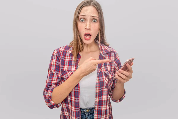 Verbaasd en ongerust vrouw staat met de mond wijd geopend. Ze houdt van telefoon in één hand en wijst op het met de vinger van een andere. Geïsoleerd op een grijze achtergrond. — Stockfoto