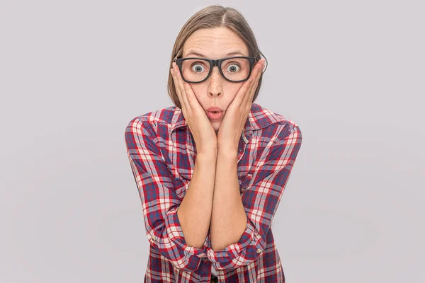 Erstaunt und aufgeregt blickt das junge Model trotz Brille in die Kamera. hält sie die Hände auf die Wangen. junge Frau wundert sich. isoliert auf grauem Hintergrund. — Stockfoto