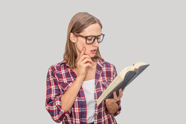 Smart and young woman reads book. She holds it opened. Model points on cheek. She wears glasses. Young woman is concentrated and serious. Isolated on grey background. — Stock Photo, Image