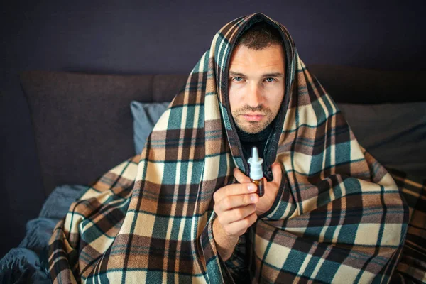 Sick young man looks on camera. He holds nose spray in hand. Guy is serious, He is completely covered with blanket. Young man is in bedroom.