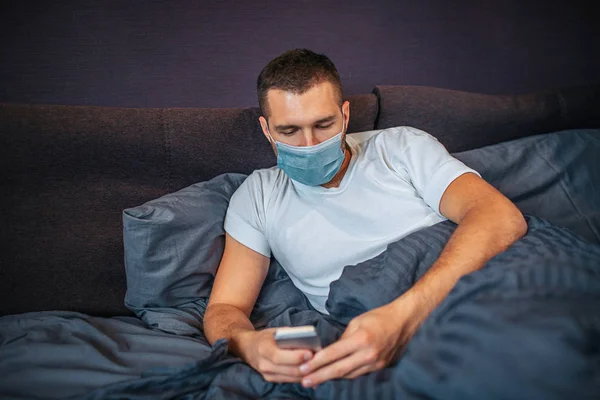 Sick young man is lying on bed and wearing mask. He holds phone in hands and look at it. Guy is calm and concentrated. Young man in in bedroom.